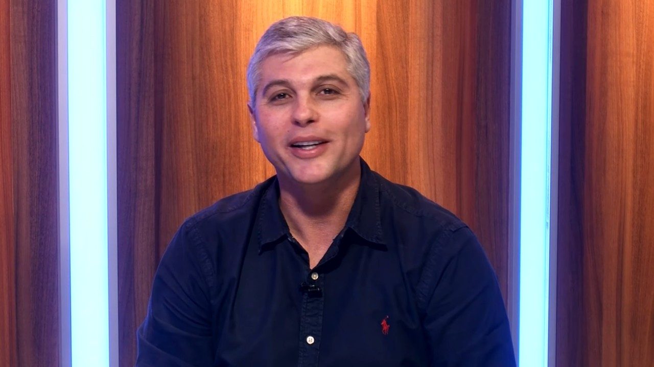 Joel Datena sorri durante participação em estúdio de TV com painel de madeira e luzes verticais ao fundo