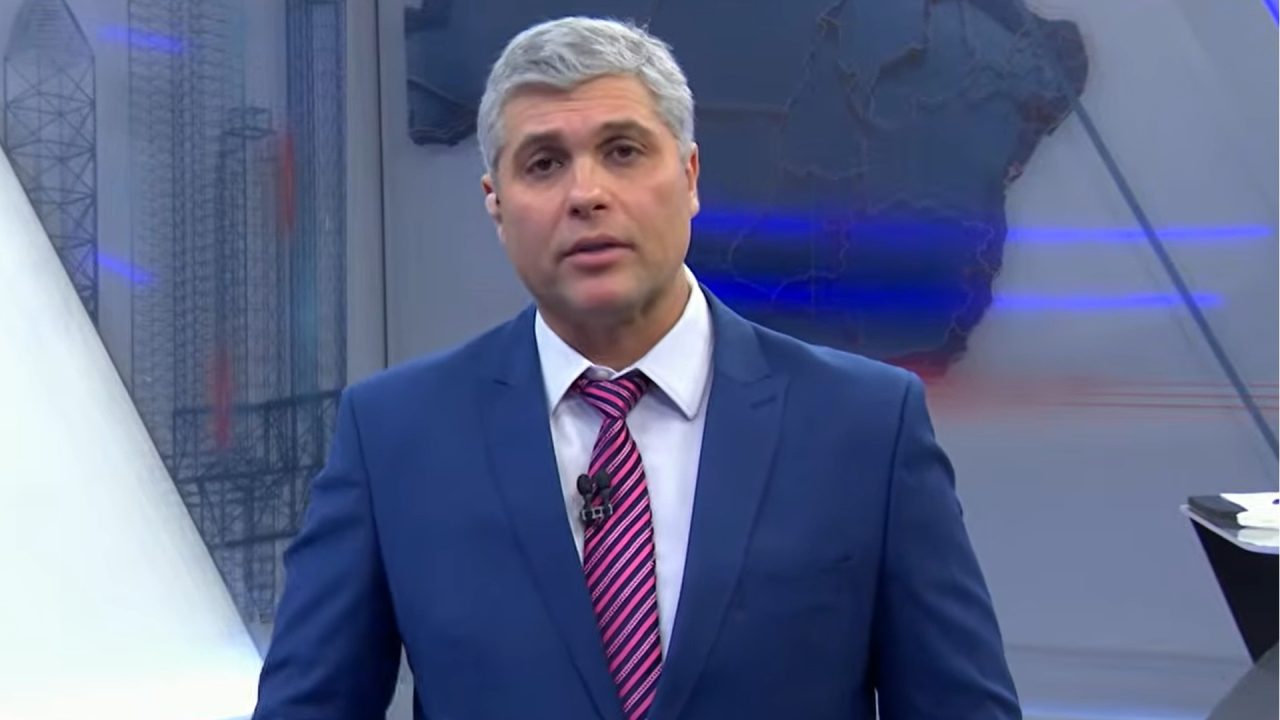 Joel Datena usa terno azul e gravata listrada durante transmissão do Brasil Urgente, em estúdio com mapa ao fundo.