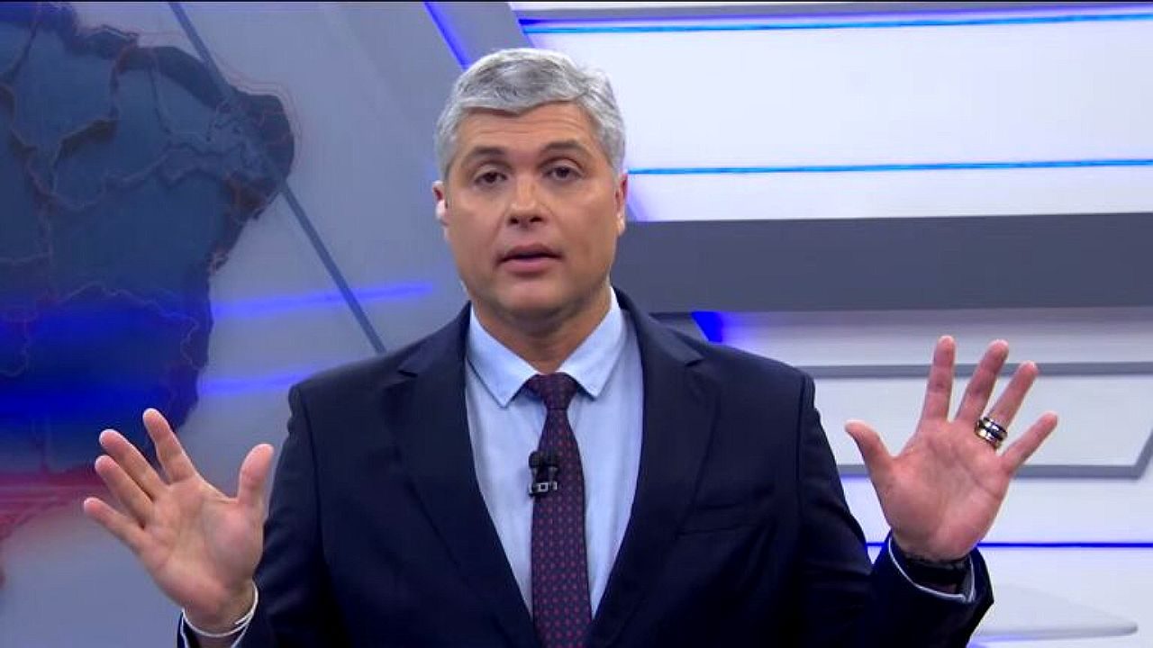 Joel Datena apresenta telejornal ao vivo com as mãos erguidas diante de painel gráfico em estúdio da Band