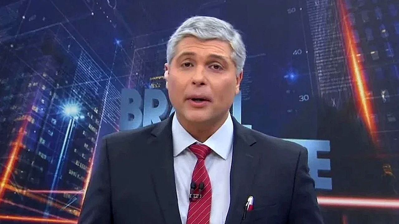Joel Datena apresenta telejornal Brasil Urgente usando terno escuro e gravata vermelha diante de cenário urbano digitalizado