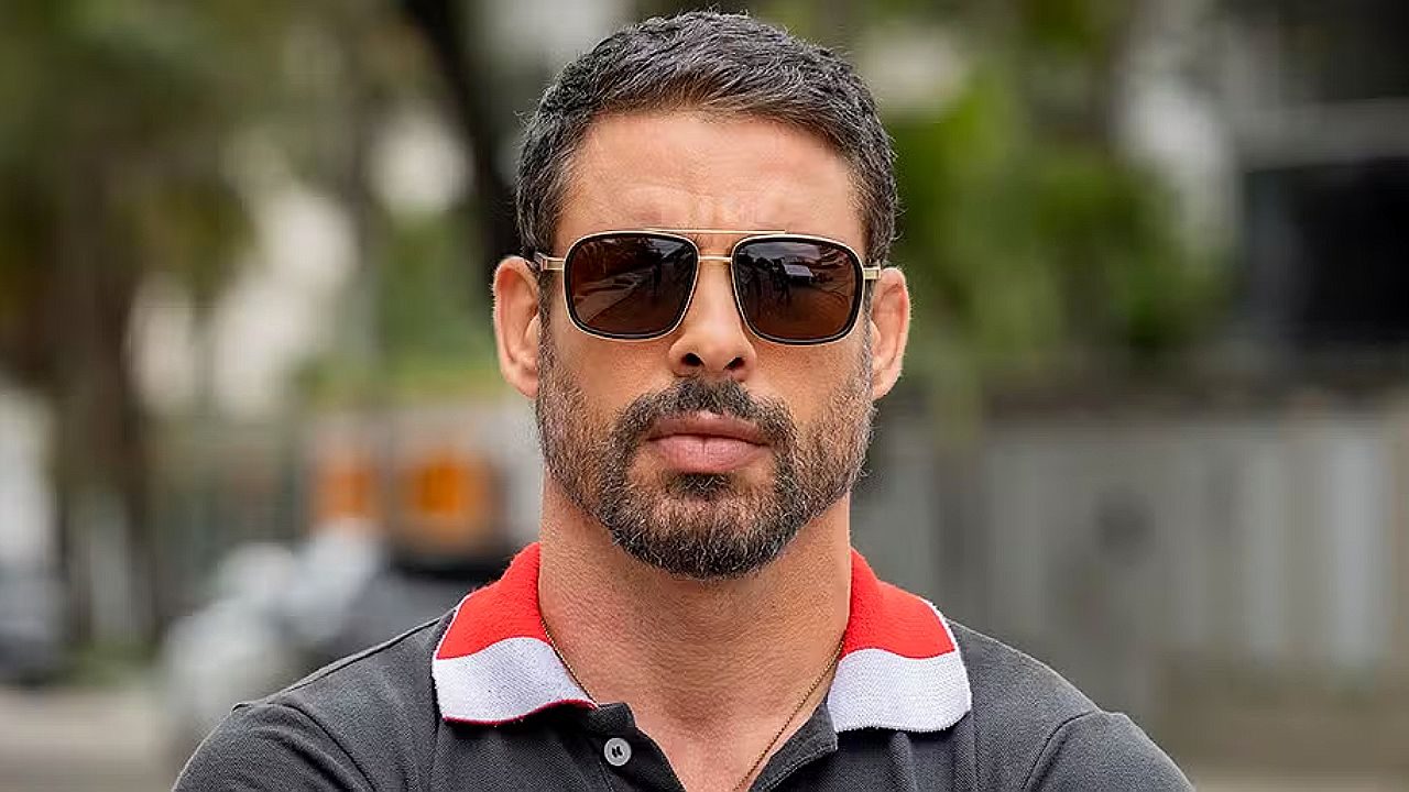 Ator de óculos escuros encara a câmera em cena da novela Jogada de Risco nas ruas da cidade