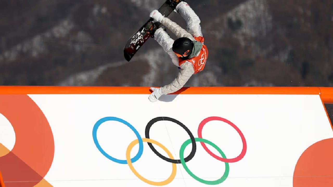 Atleta realiza manobra de snowboard durante competição de inverno, deslizando sobre estrutura com o símbolo olímpico impresso, em cenário de montanhas ao fundo.