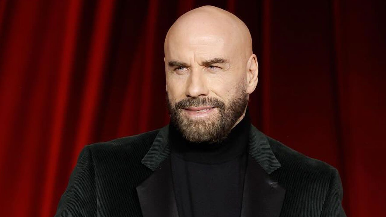 John Travolta com blazer escuro e fundo de cortina vermelha durante evento ou premiação