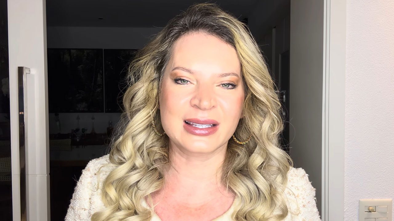 Joice Hasselmann de cabelos loiros ondulados, brincos de argola dourados e casaco claro, olhando para a câmera e falando em ambiente interno iluminado