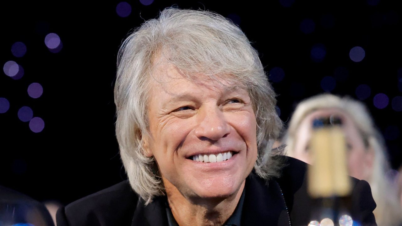 Homem de cabelo grisalho sorri em evento noturno com luzes desfocadas ao fundo