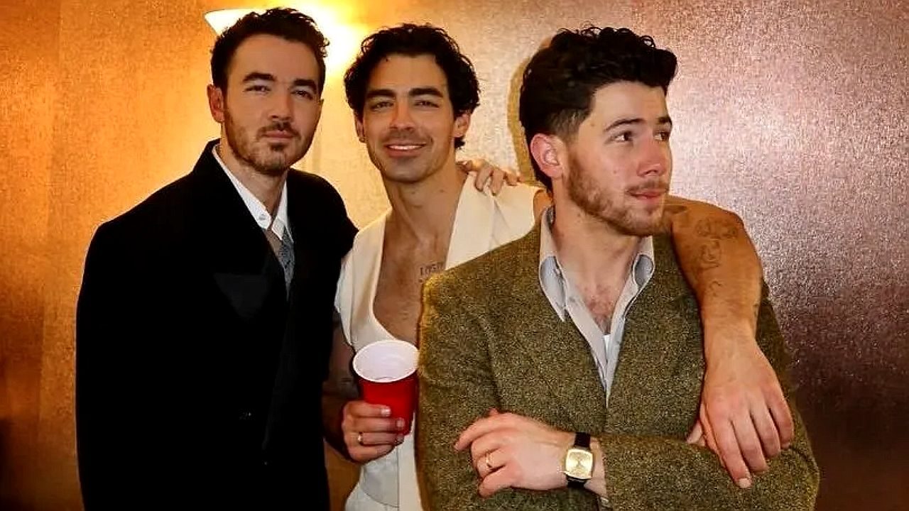 Integrantes dos Jonas Brothers posam juntos em bastidores de evento com copo vermelho nas mãos