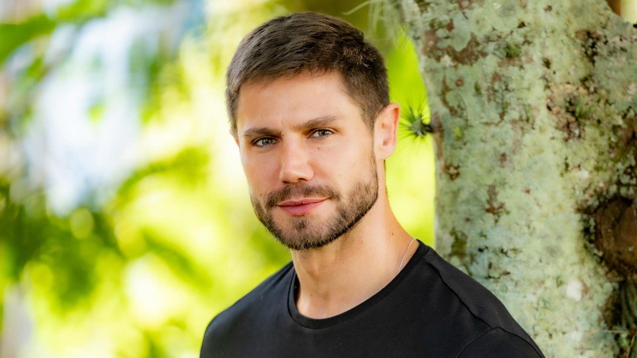 Jonas Sulzbach posa ao ar livre encostado em árvore usando camiseta preta com expressão séria e olhar direto para a câmera