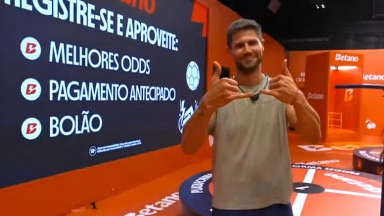 Jonas Sulzbach sorri e faz gesto com as mãos diante de painel da Betano durante ação comercial no BBB 26