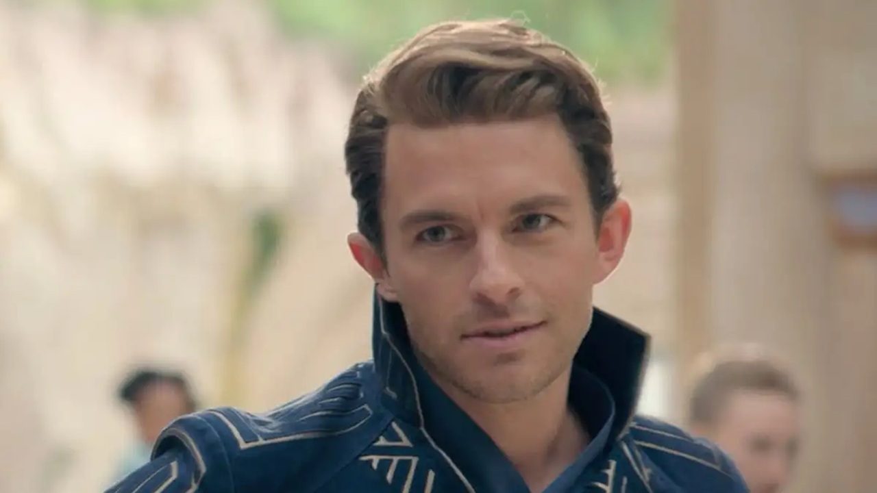 Homem de roupa azul com gola alta olha para frente em ambiente claro de época ou fantasia