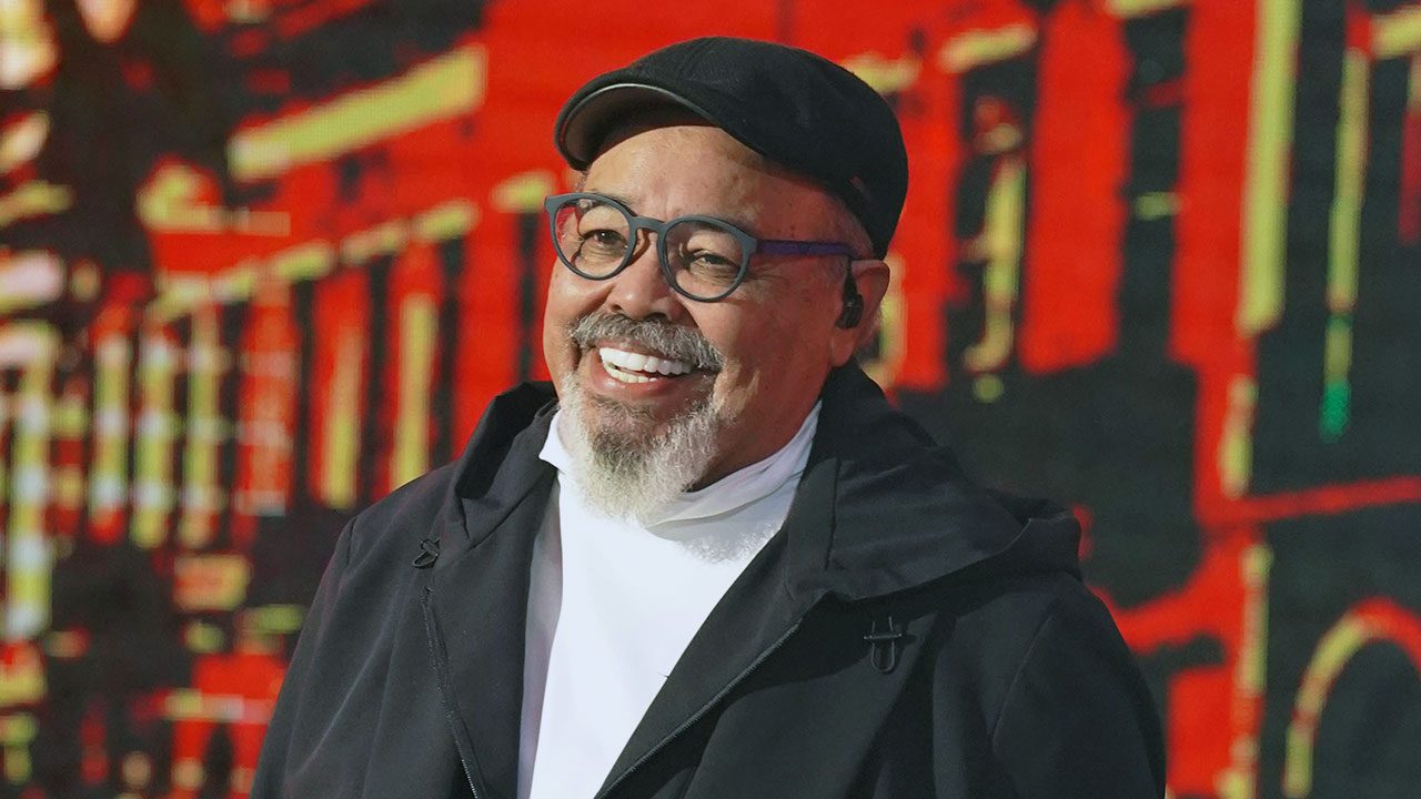 Cantor Jorge Aragão sorridente, com cavanhaque branco, óculos de aro escuro e boné preto, em show em palco com LED vermelho