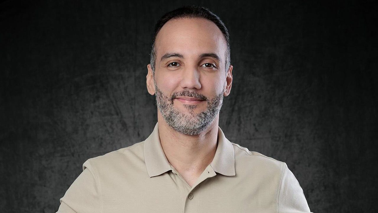 Jorge Iggor com leve sorriso usando camisa polo bege diante de fundo escuro