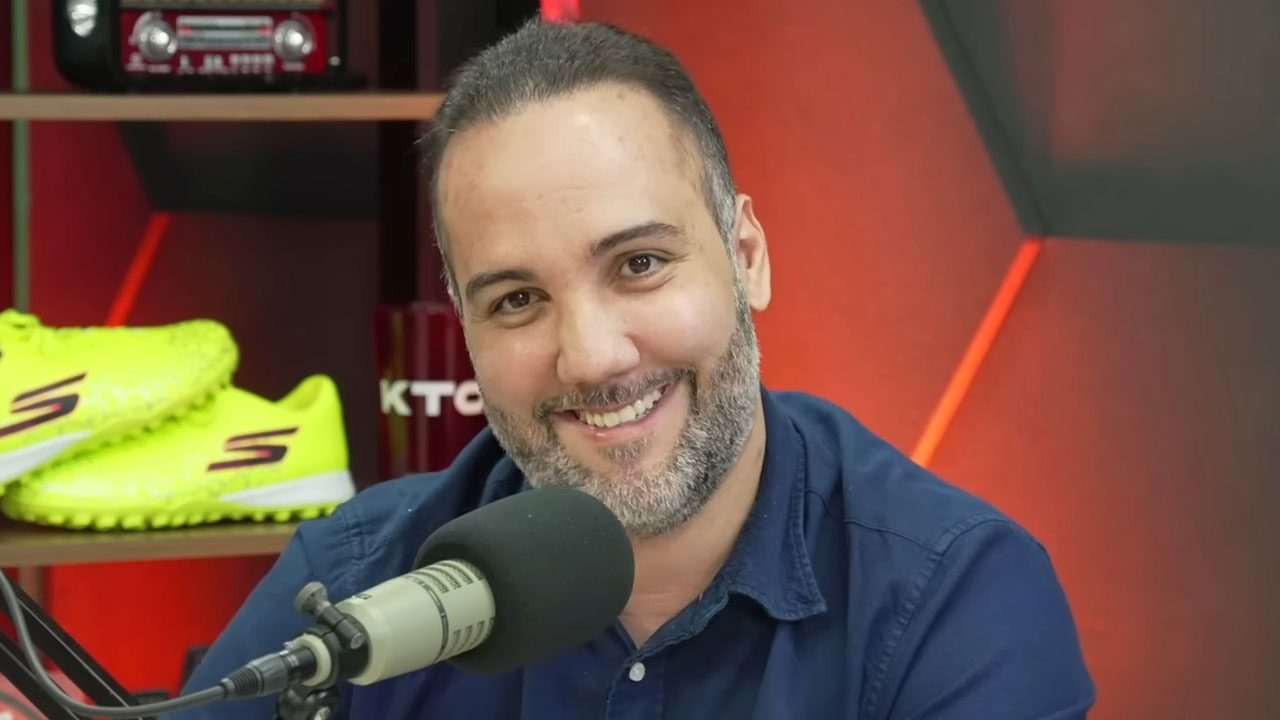 Jorge Iggor sorri diante de microfone durante podcast com cenário vermelho ao fundo