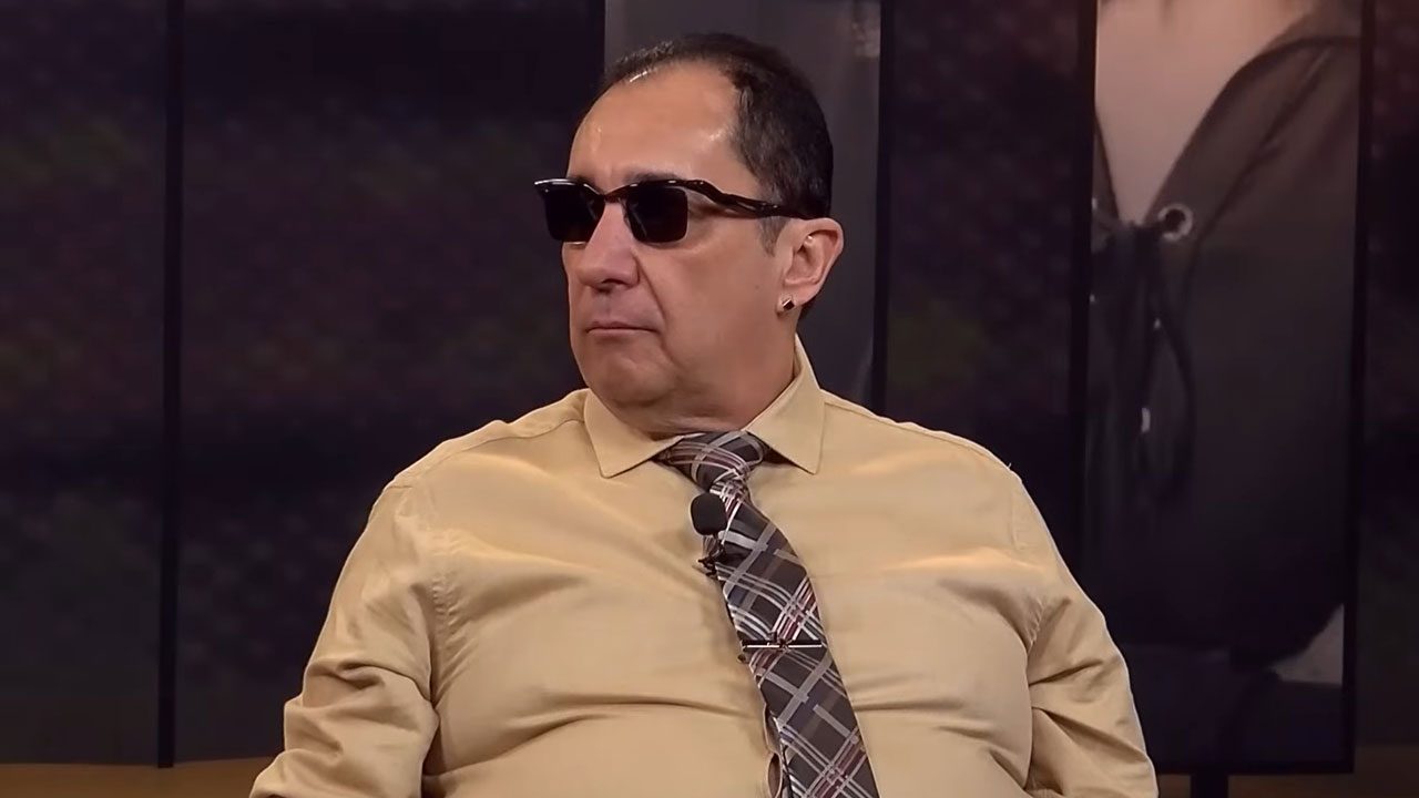 Jorge Kajuru sentado em estúdio de televisão, usando óculos escuros, camisa bege e gravata estampada
