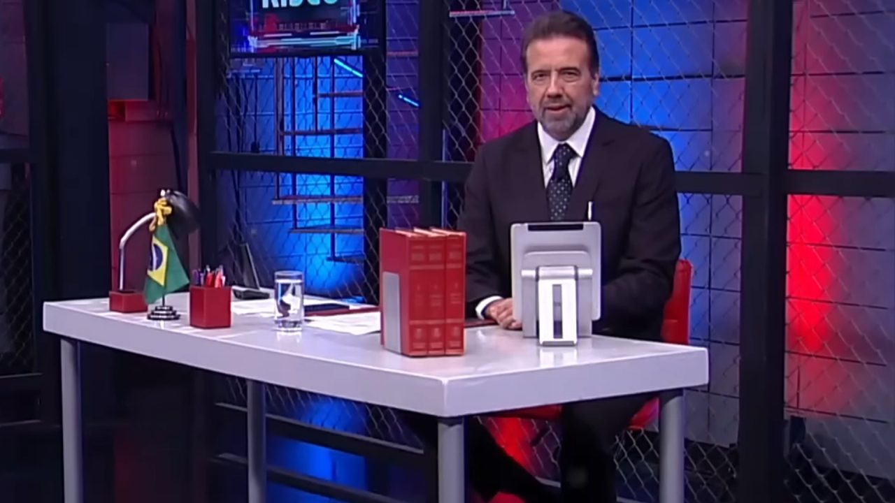Apresentador Jorge Lordello sentado à mesa em estúdio da RedeTV! durante gravação de programa, com cenário iluminado em azul e vermelho.