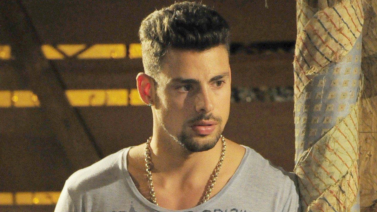 Cauã Reymond como Jorginho observa com expressão séria em cena da novela Avenida Brasil em ambiente interno rústico