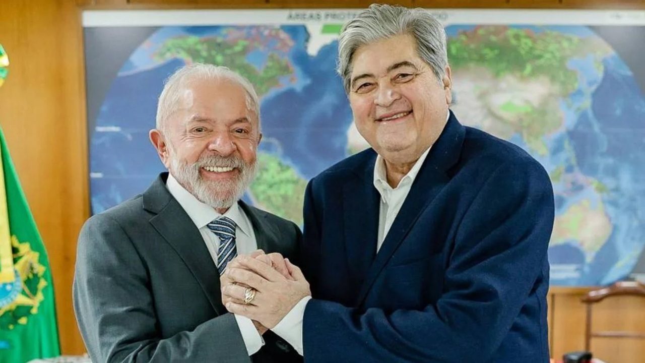 Dois homens de terno sorriem e apertam as mãos em gabinete com mapa-múndi grande na parede ao fundo e bandeira do Brasil à esquerda, um deles com barba branca e cabelo grisalho vestindo terno cinza e gravata azul, o outro de cabelo grisalho penteado para o lado usando terno azul escuro sem gravata, ambos posicionados em frente a mesa de madeira em ambiente institucional bem iluminado