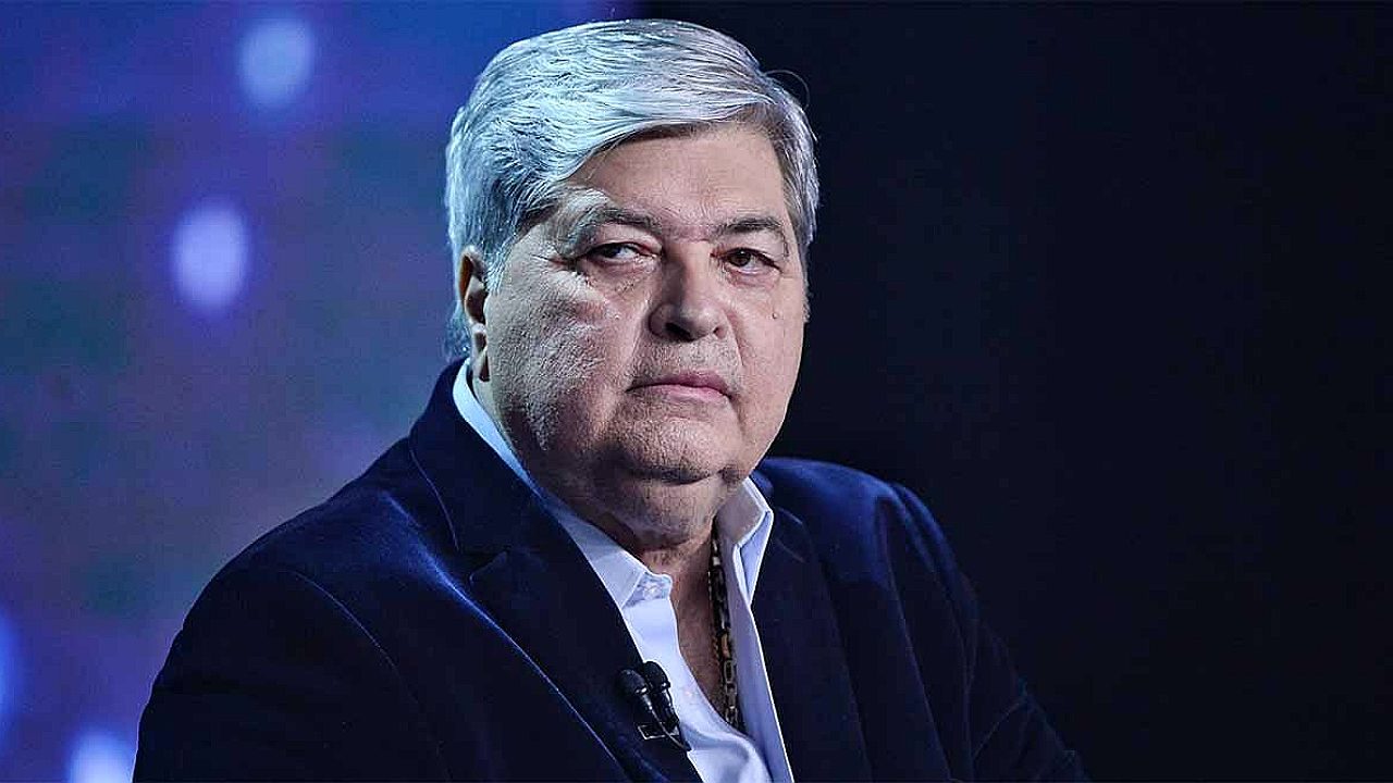 Apresentador Datena de terno escuro e camisa branca encara a câmera durante participação em programa de televisão com iluminação azul ao fundo