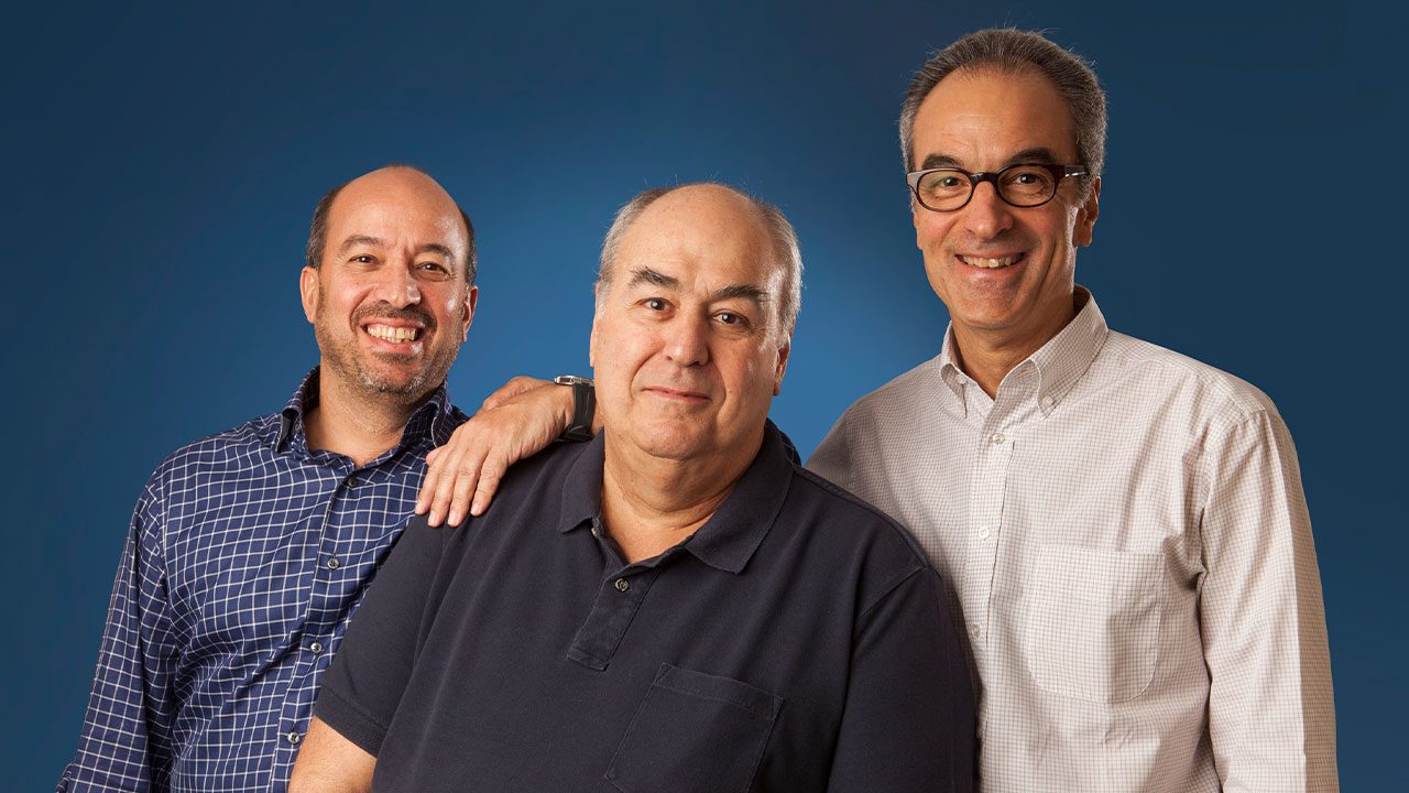 Acionistas da Globo, José Roberto Marinho, Roberto Irineu Marinho e João Roberto Marinho posando juntos em frente a fundo azul