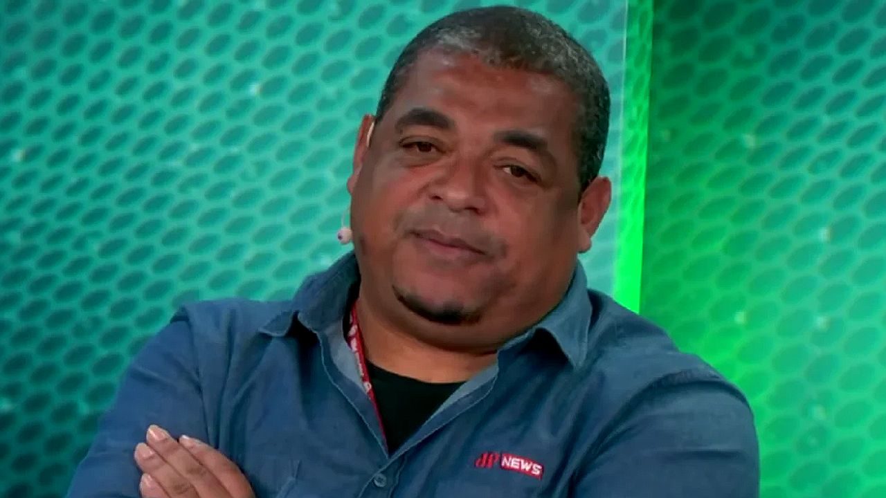 Homem de camisa azul com logotipo da Jovem Pan News e microfone de lapela participa de programa esportivo em estúdio com cenário verde ao fundo
