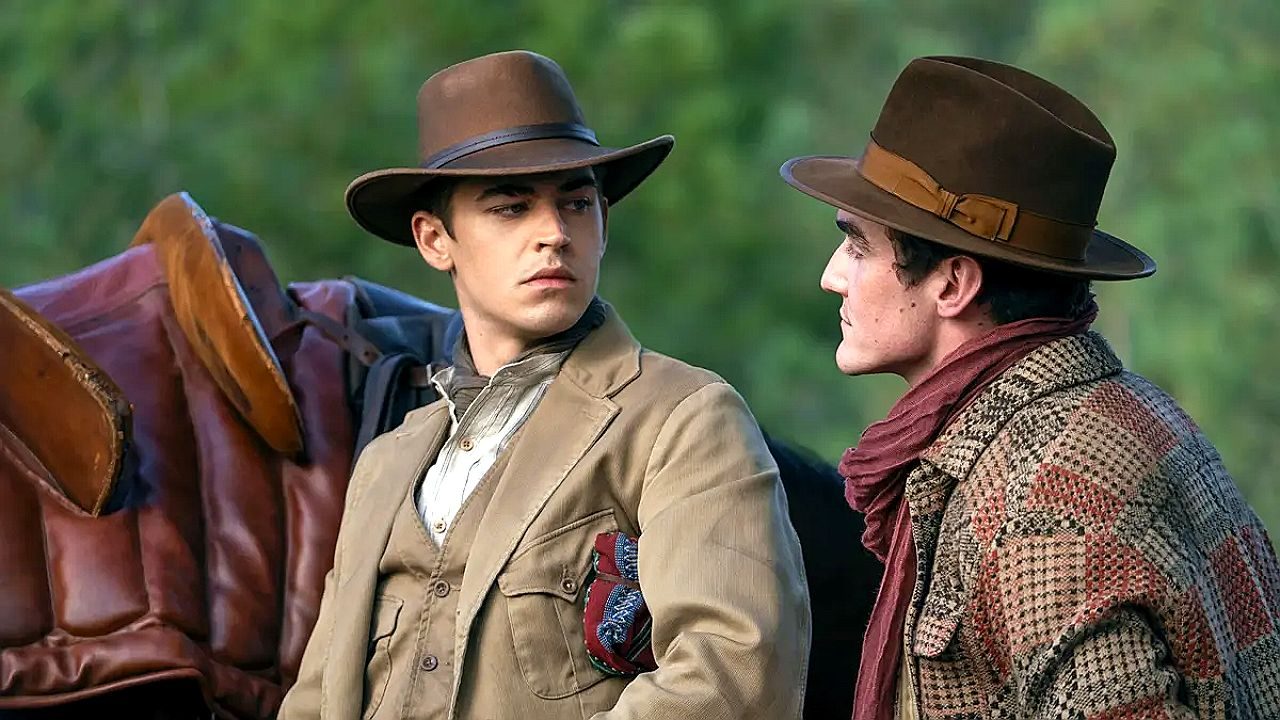 Dois homens com chapéu e roupas de época conversam ao lado de sela de cavalo em cena de faroeste