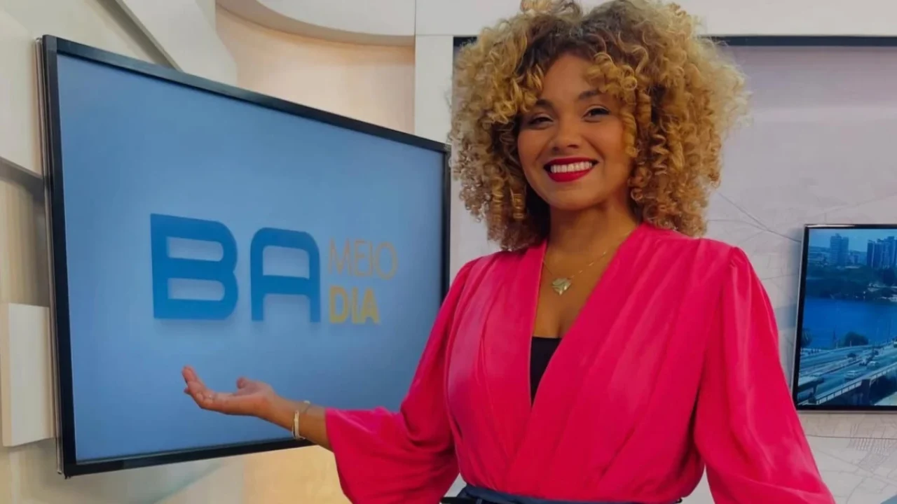 A imagem mostra uma apresentadora sorridente em um estúdio de telejornal. Ela está ao lado de uma tela que exibe o logotipo “BA Meio Dia”. A mulher usa um vestido rosa vibrante e tem cabelo cacheado e volumoso. O cenário é moderno e bem iluminado, com tons claros e elementos visuais de um ambiente televisivo profissional