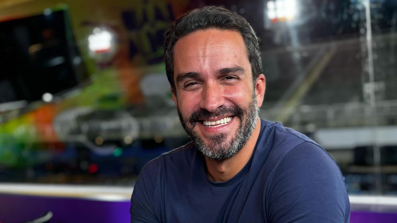 JP Vergueiro sorrindo com camiseta azul em ambiente interno com luzes e painel colorido ao fundo