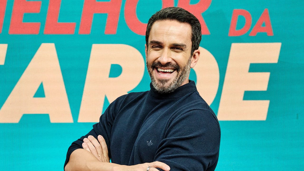 JP Vergueiro sorri de braços cruzados em frente ao telão com logo colorido do programa Melhor da Tarde