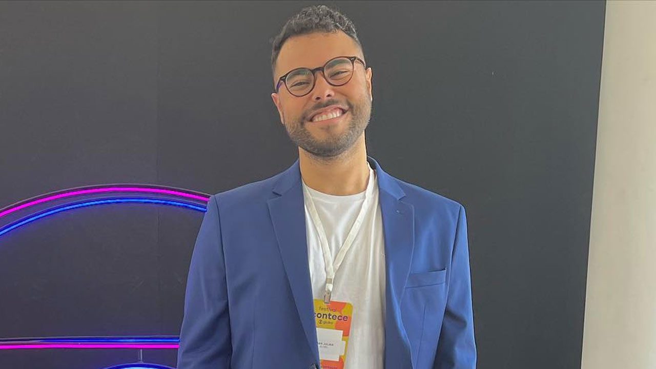 Juan Jullian, de óculos, camiseta branca e blazer azul, sorri usando um crachá de evento em frente a um painel escuro com luzes neon