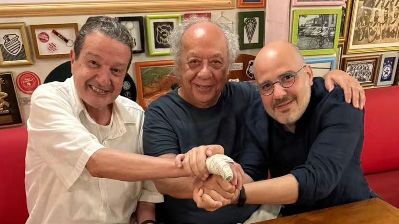Juca Kfouri, José Trajano e André Basbaum sorriem e unem as mãos em restaurante com quadros de futebol ao fundo