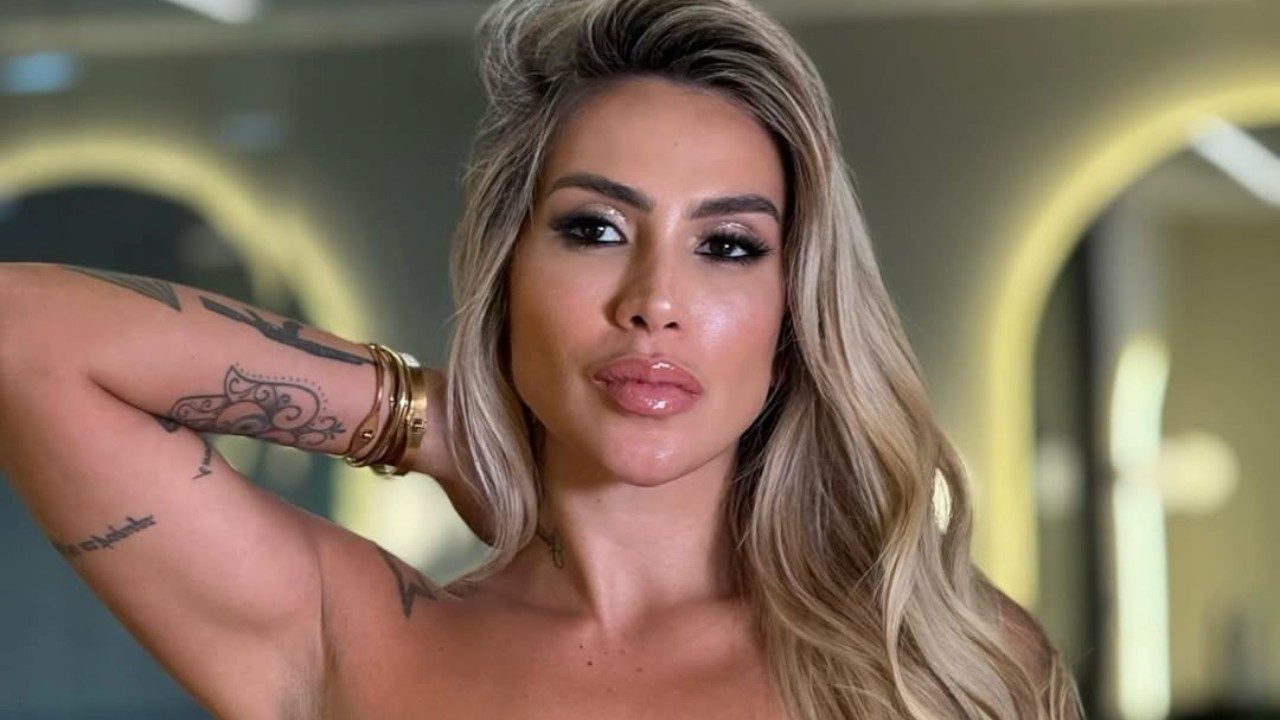 Juju Ferrari posa com a mão no cabelo exibindo tatuagens no braço em ambiente interno com iluminação suave ao fundo