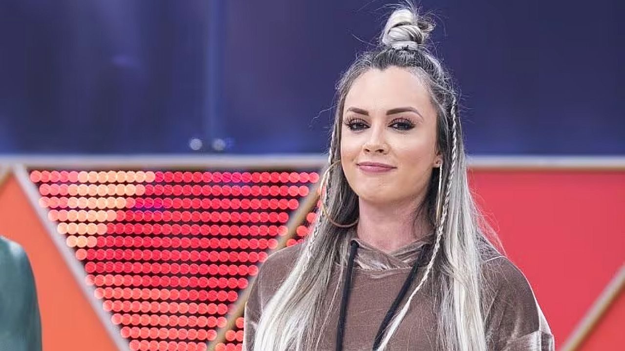 Mulher de cabelo longo preso em coque alto participa de programa de televisão em estúdio com painel de luzes vermelhas ao fundo