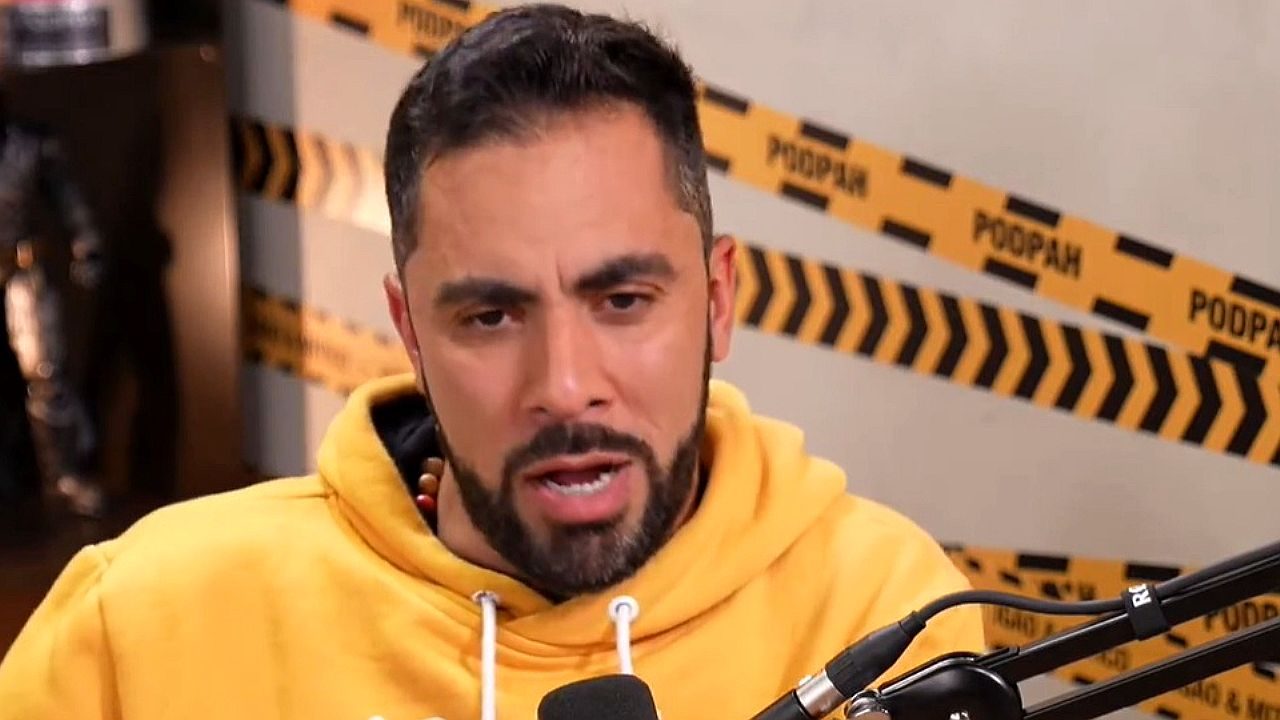 Homem de moletom amarelo fala ao microfone em estúdio de podcast com fundo decorado com fitas de isolamento