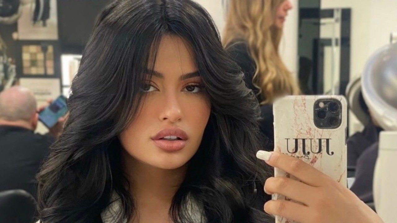 Júlia Medeiros tira selfie no espelho em salão de beleza com cabelo volumoso e celular na mão enquanto pessoas aparecem ao fundo