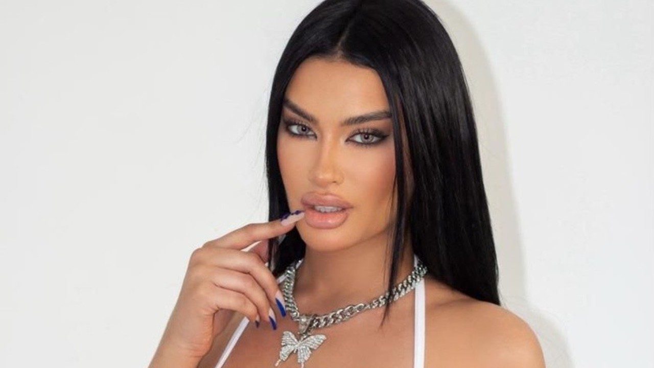 Júlia Medeiros com cabelo preto liso toca os lábios com a mão usando top branco e colar prateado diante de fundo claro