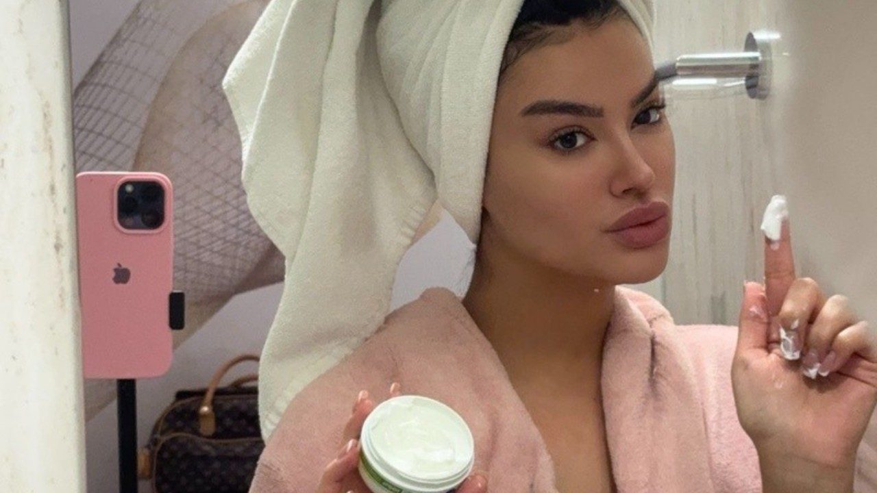 Júlia Medeiros posa em selfie no espelho com toalha na cabeça e roupão rosa segurando pote de creme e aplicando produto no dedo em banheiro