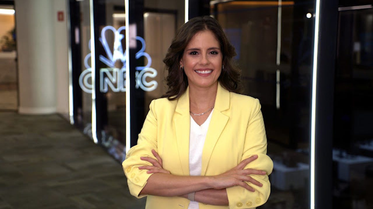 Juliana Munaro sorri com os braços cruzados, usando um blazer amarelo claro e camiseta branca, em frente a uma parede de vidro com o logotipo iluminado da CNBC ao fundo