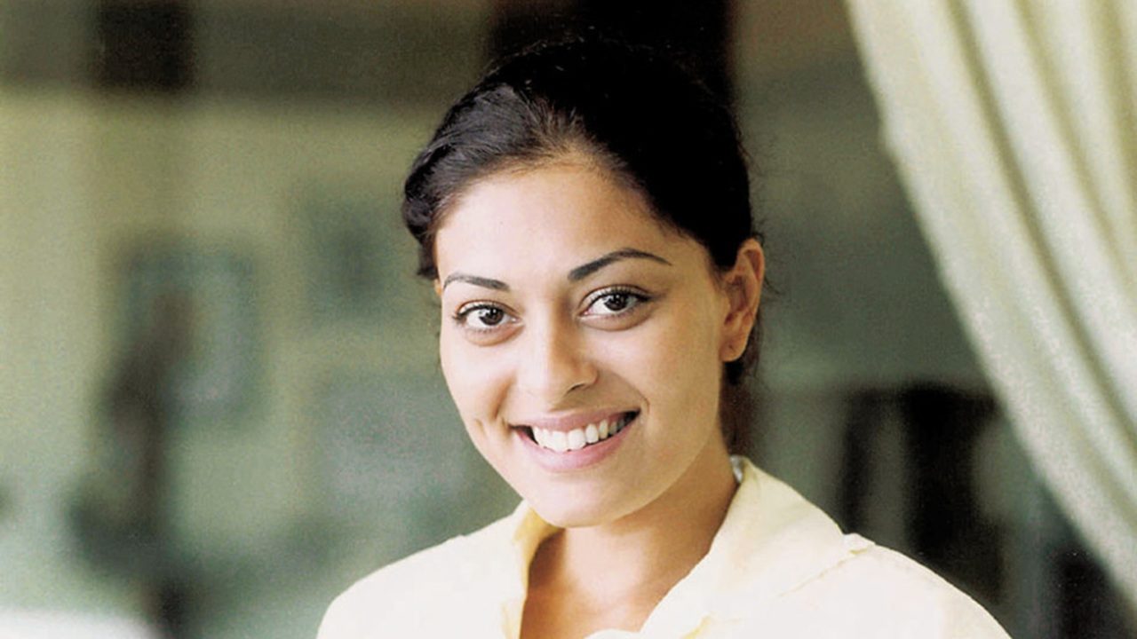 Mulher jovem sorrindo em um retrato iluminado de forma suave. Ela tem cabelos escuros presos para trás, covinhas discretas e veste uma camisa clara. O fundo desfocado sugere um ambiente interno, transmitindo leveza e naturalidade.