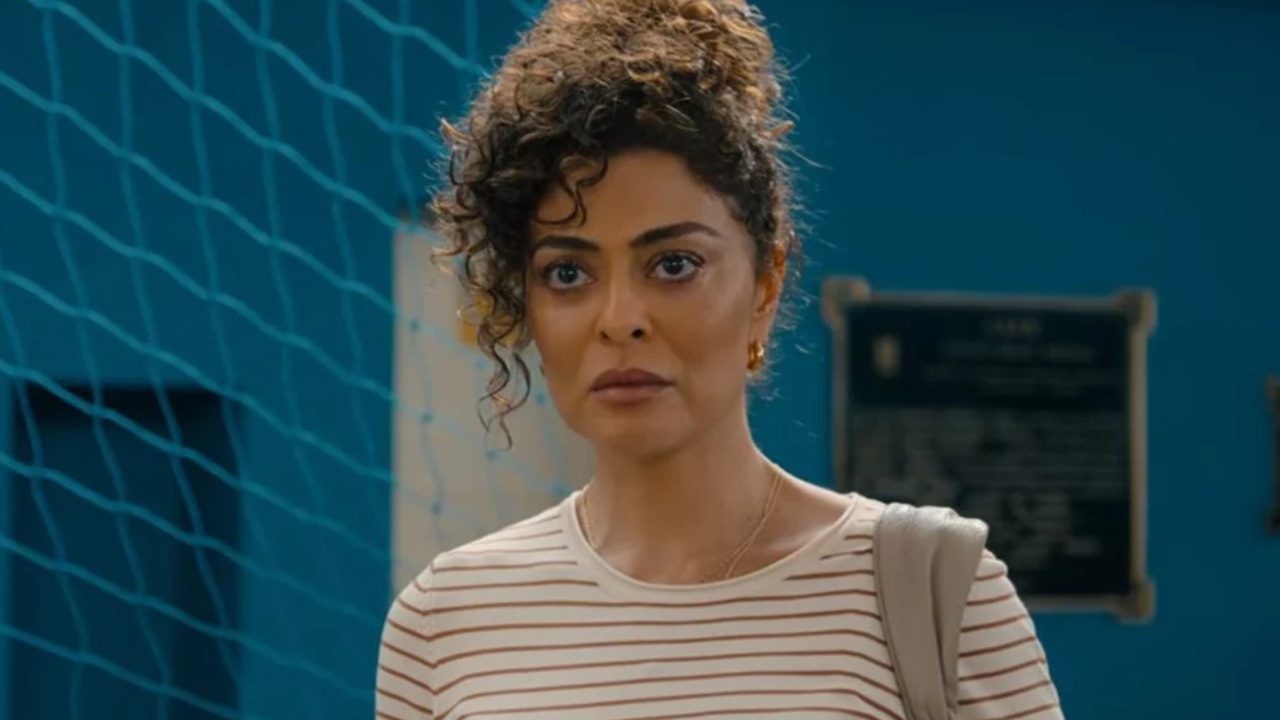 A imagem mostra uma mulher de expressão séria, com cabelos cacheados presos no alto da cabeça e alguns fios soltos. Ela usa uma blusa listrada em tons claros e carrega uma bolsa no ombro. O fundo azul e as placas desfocadas sugerem que a cena acontece em um ambiente público, como uma escola ou instituição