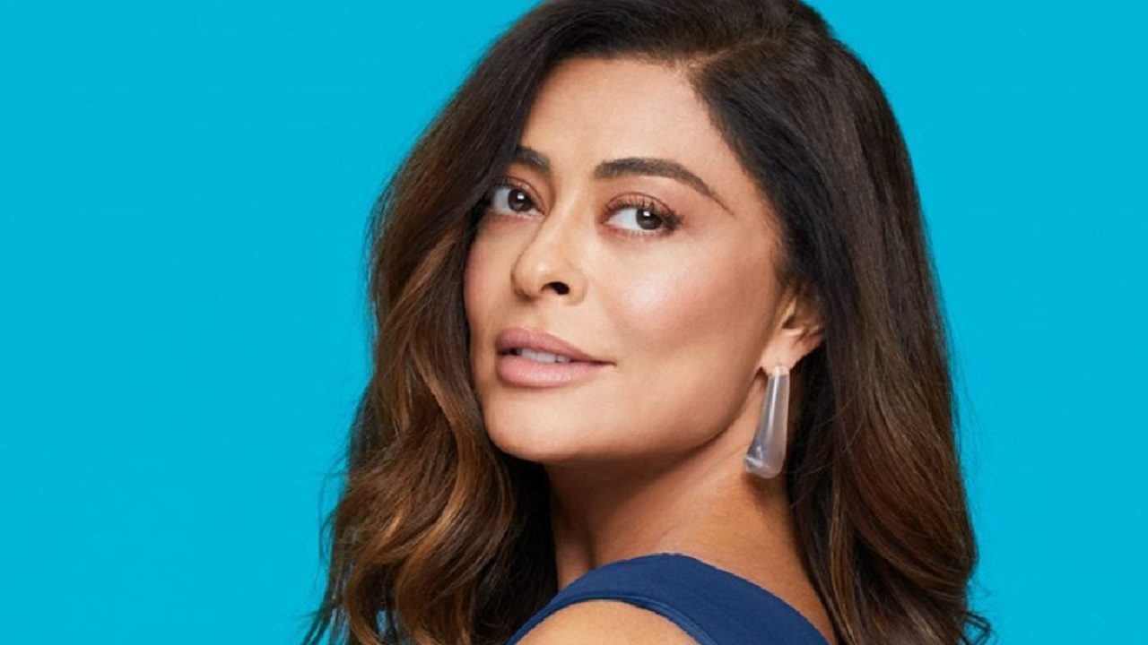 Mulher de cabelos castanhos ondulados, usando vestido azul e brincos prateados, posa levemente de lado diante de um fundo azul-claro