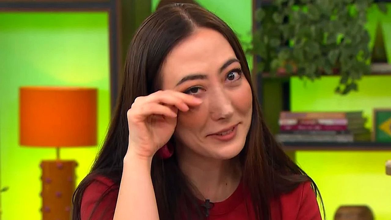 Mulher de blusa vermelha enxuga lágrima durante entrevista em estúdio com cenário colorido ao fundo