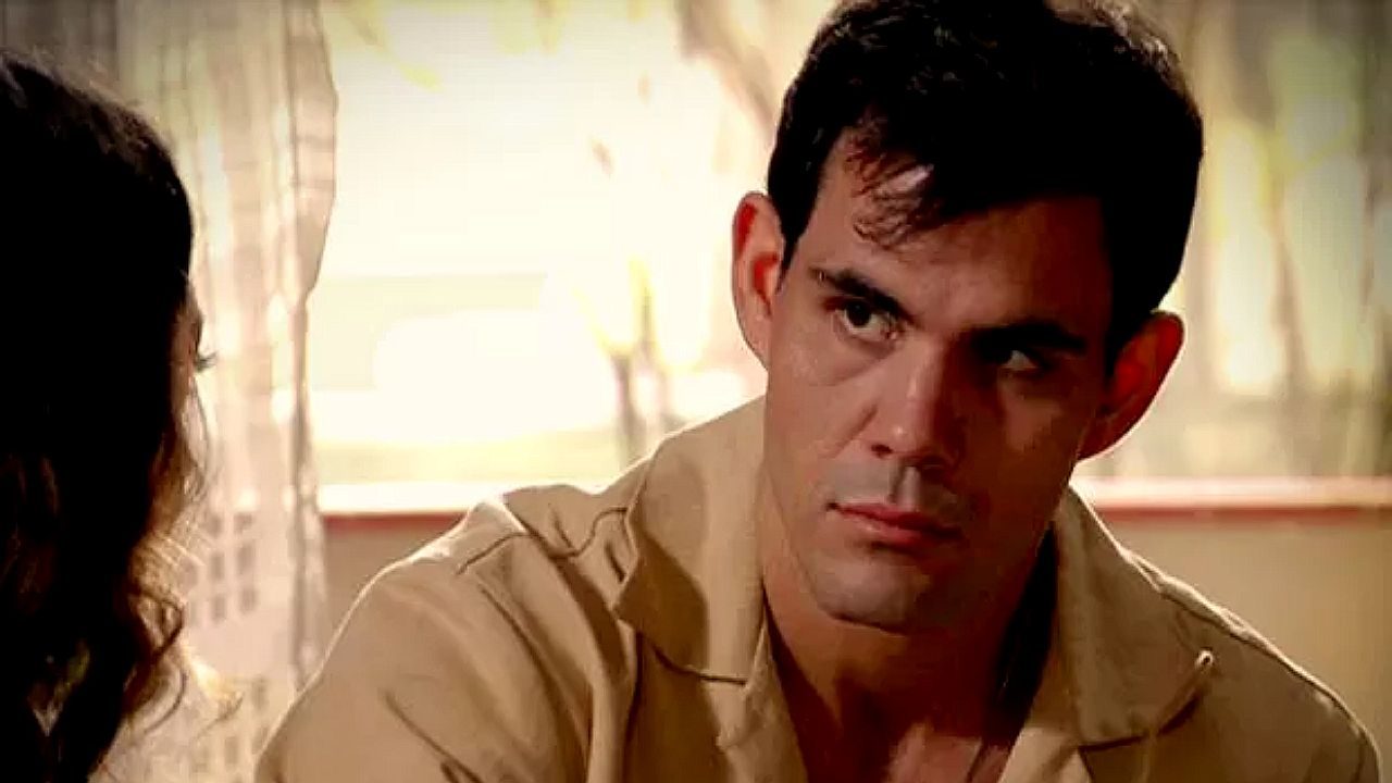 Juliano Cazarré de camisa bege encara mulher com expressão séria em cena de novela ambientada em sala iluminada