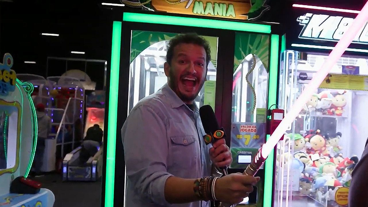 Homem segura microfone e sabre de brinquedo em arcade diante de máquina de pelúcias iluminada