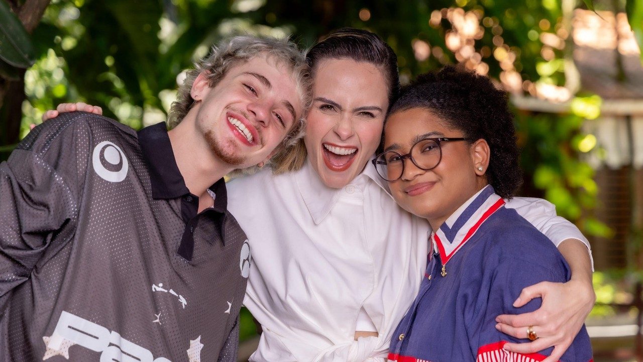 Juliano Floss, Ana Paula Renault e Milena abraçados e sorrindo juntos em área externa com vegetação ao fundo