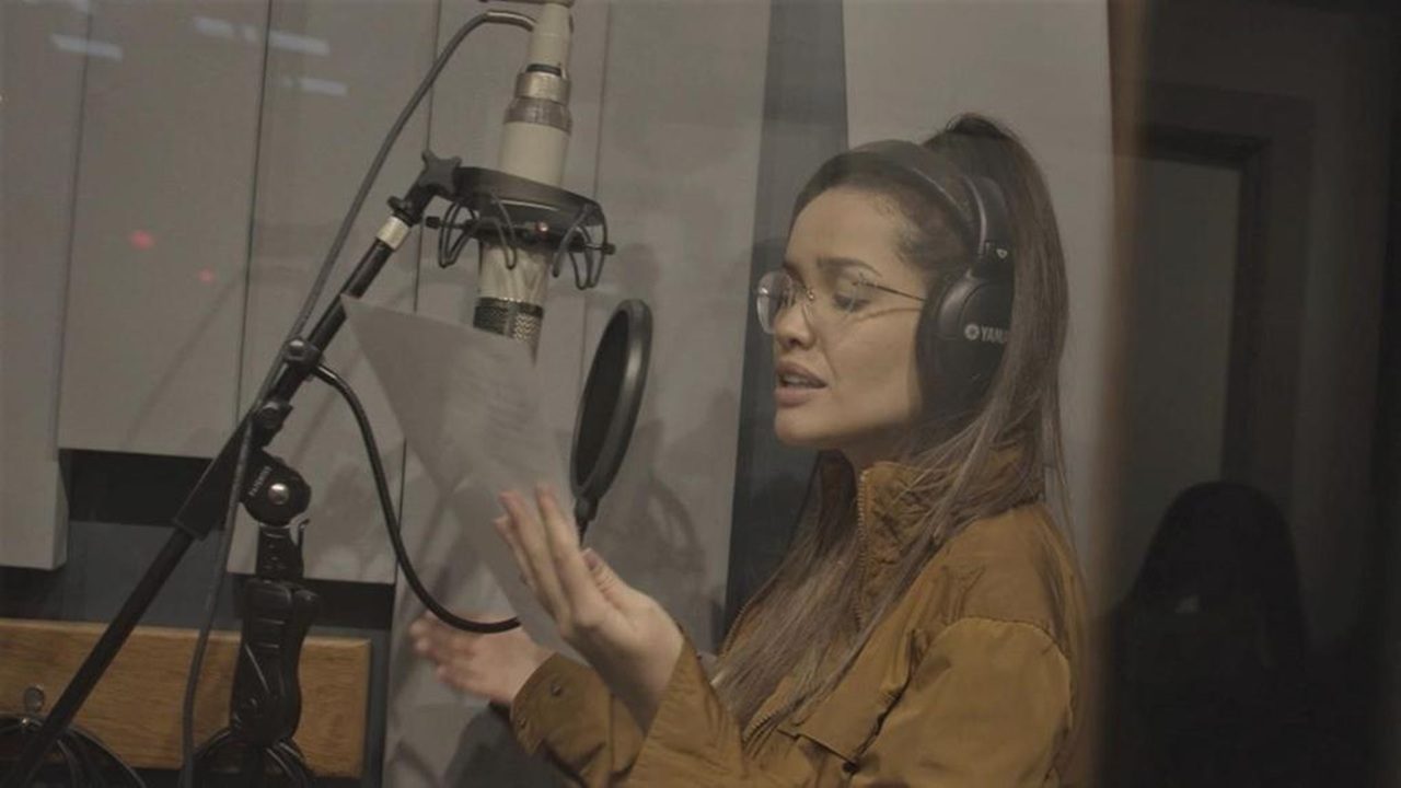 Mulher em estúdio de gravação, usando fones de ouvido grandes e óculos, canta ou narra enquanto segura uma folha de papel diante de um microfone profissional suspenso. O ambiente é iluminado de forma suave, com paredes acústicas ao fundo.