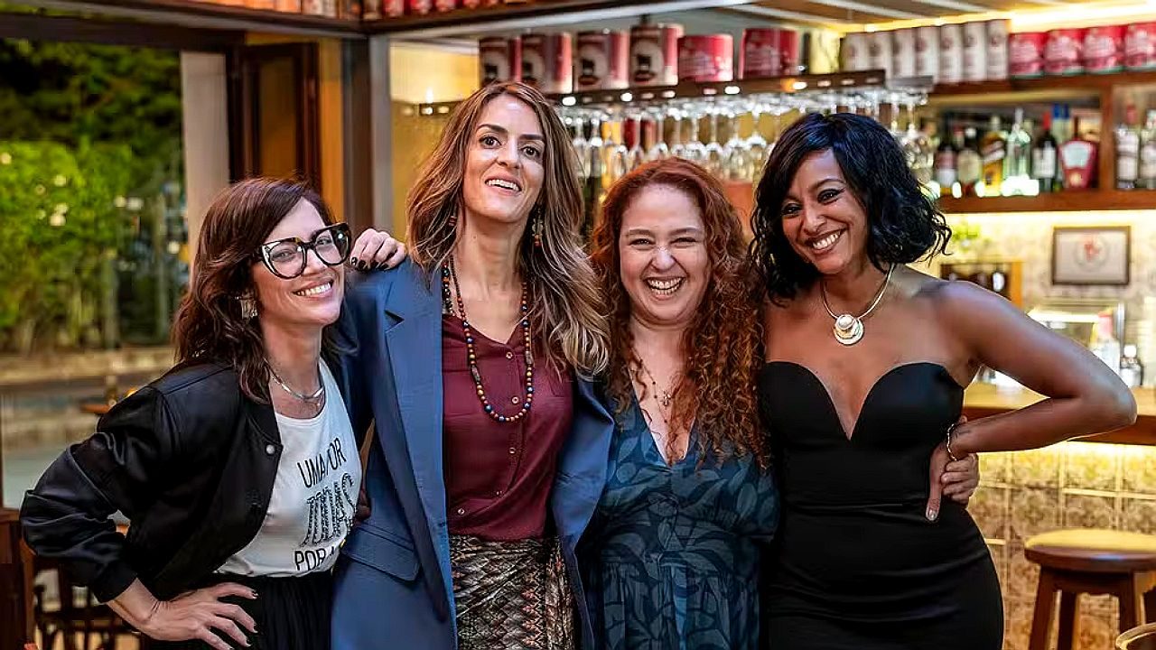 Quatro mulheres estão posando juntas e sorrindo em frente ao balcão de um bar ou restaurante. Elas estão próximas umas das outras, com os braços apoiados ou abraçados. Ao fundo aparecem prateleiras com garrafas, copos e utensílios de bar. Cada uma veste roupas diferentes, variando entre casacos, vestidos e blusas elegantes, sugerindo um encontro social ou cena em ambiente noturno.