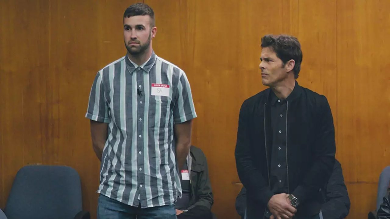 Dois homens em pé em uma sala de madeira, um usando camisa listrada com crachá de jurado e o outro de jaqueta escura, parecem participar de uma audiência ou julgamento.