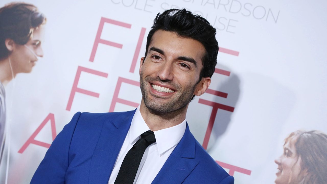 A imagem mostra um homem sorridente vestindo um terno azul, camisa branca e gravata preta, posando em frente a um fundo de evento com o título de um filme parcialmente visível