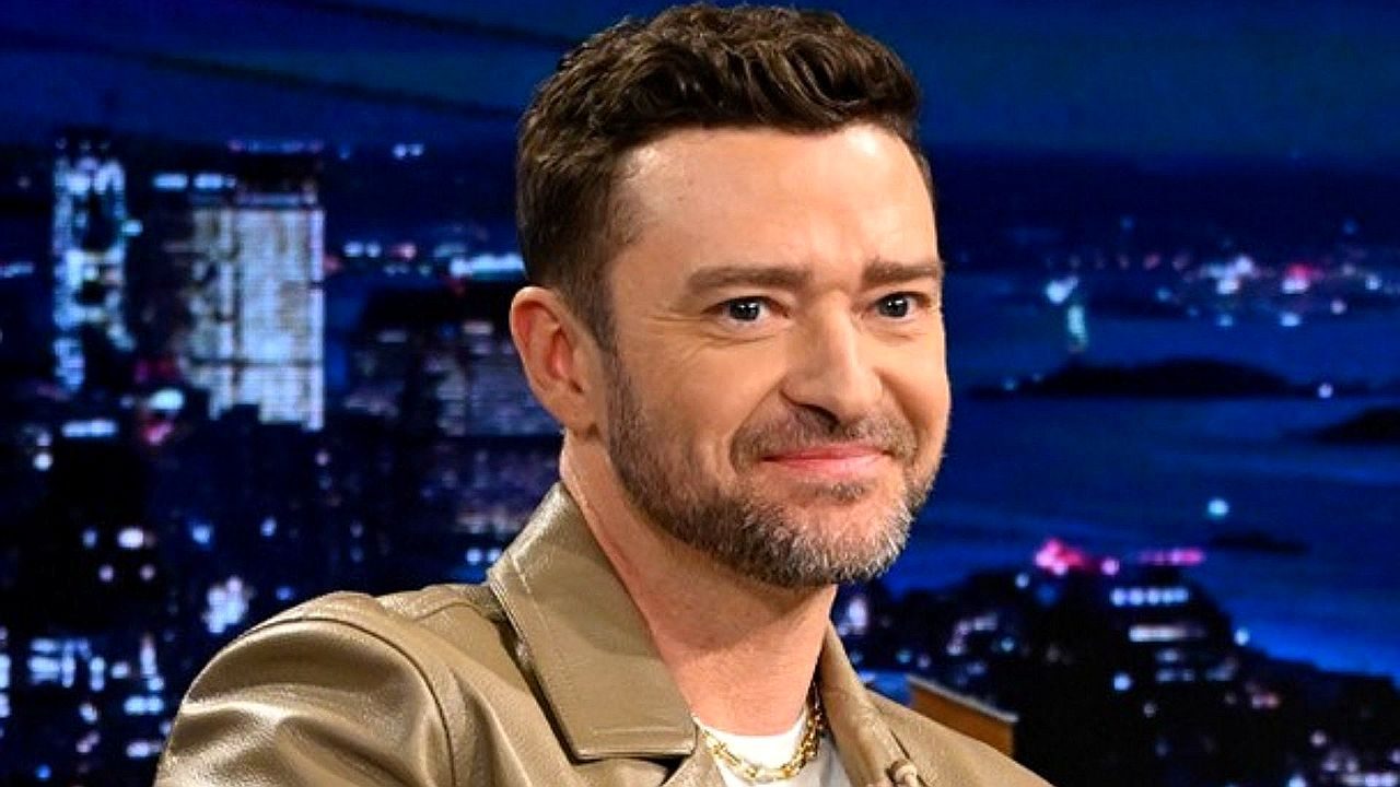 Homem sorri durante participação em talk show com cidade iluminada ao fundo no cenário do estúdio