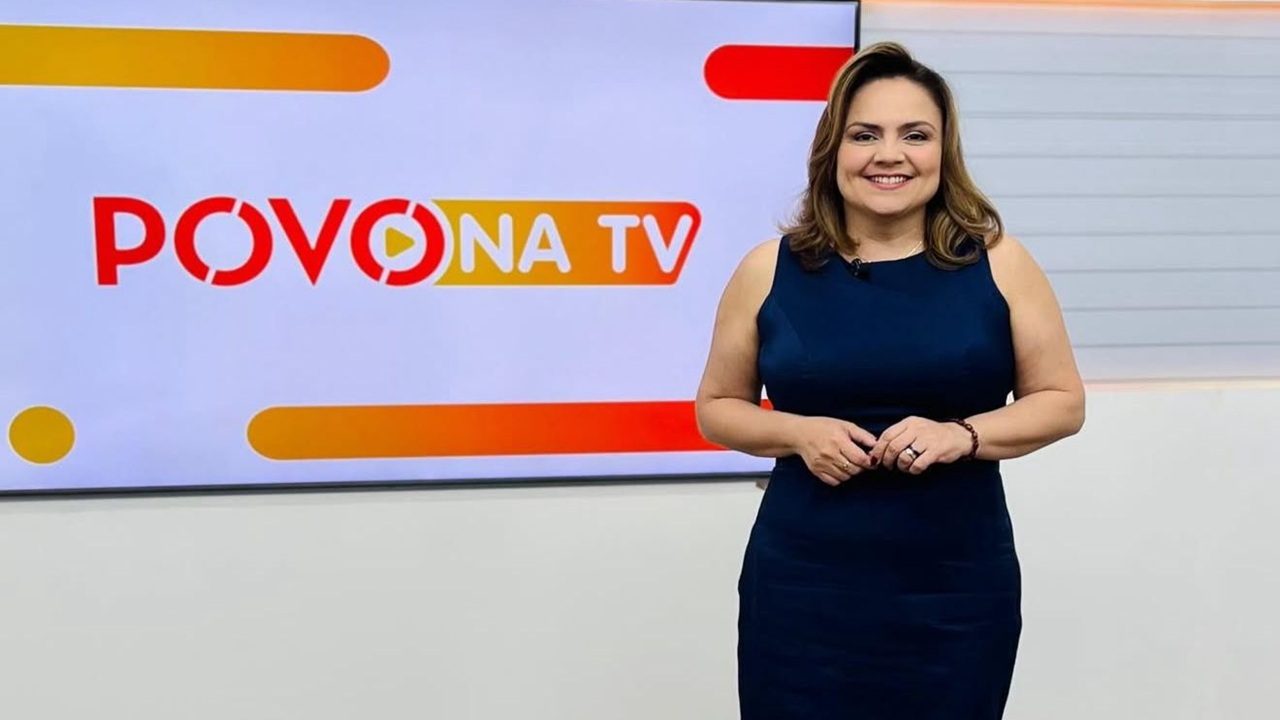 Apresentadora de vestido azul sorri em estúdio diante do telão com o logo do programa Povo na TV