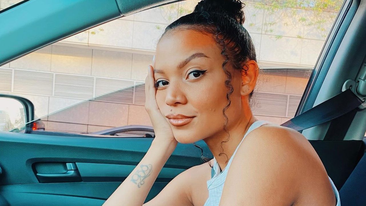 Kamila Amorim sentada no banco do carro apoiando o rosto na mão e usando blusa clara com cinto de segurança afivelado